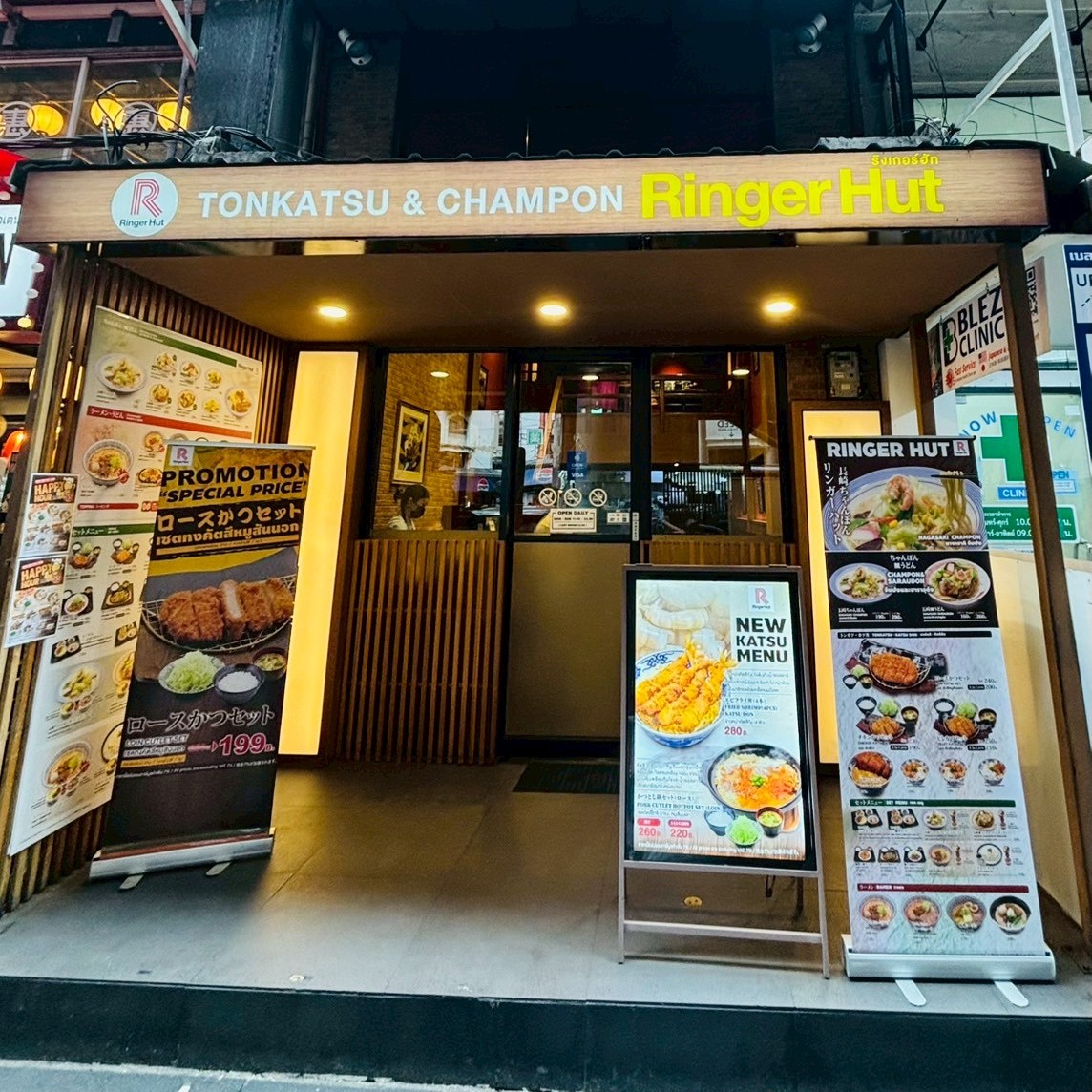 The exterior storefront of Ringer Hut Phrom Phong branch along Sukhumvit Road in Bangkok, displaying banners for TONKATSU & CHAMPON and specific promotions like the Tonkatsu set for 199 Baht. Photographed by Kaori Kondo.
