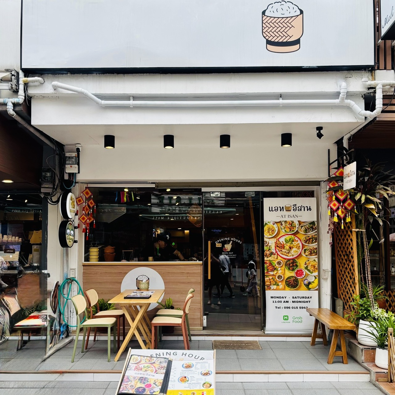 A straight-on exterior photograph of the clean, modern storefront of AT ISAN restaurant in Bangkok. It features a bright white facade with a minimalist logo of a traditional sticky rice basket, glass glass doors, outdoor wooden tables and chairs, and a vertical promotional banner showing colorful Isan dishes with opening hours. Reference filename: 11.jpg.