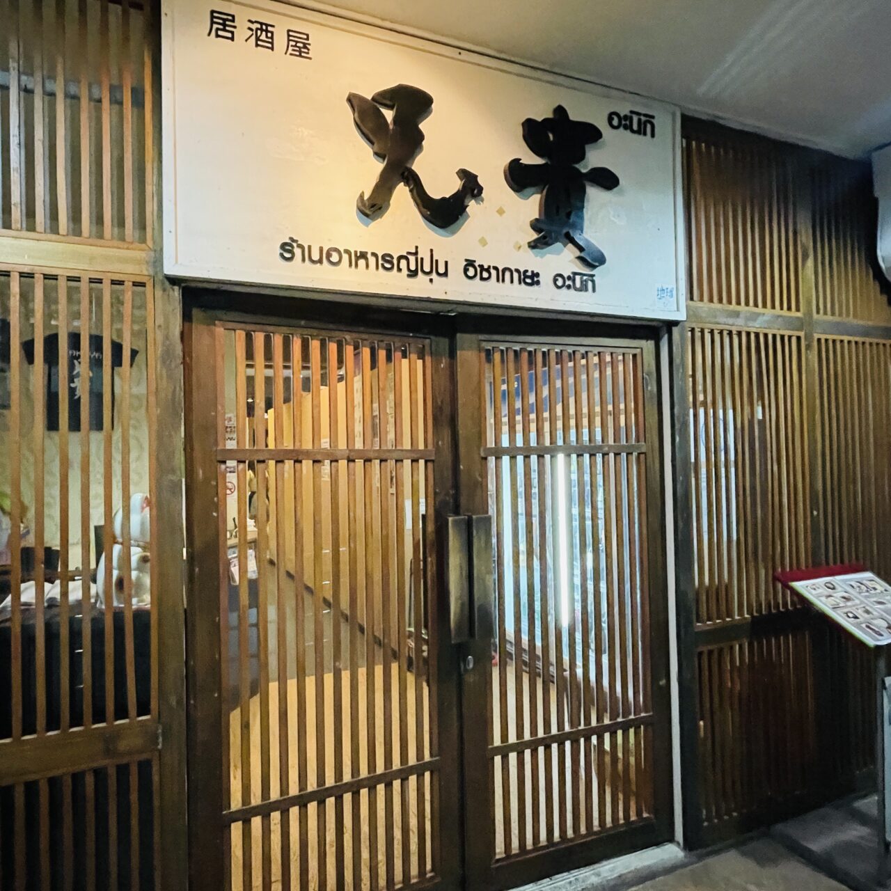 A nighttime photograph capturing the traditional Japanese-style wooden grid entrance and main signage of Izakaya Aniki in Thonglor, Bangkok. The weathered white sign features the restaurant name 'Aniki' written in large black Japanese kanji '兄貴' and Thai script 'อะนิกิ'. Below, it states 'Japanese Restaurant Izakaya Aniki' in Thai. The wooden latticework doors and shoji-style panels evoke a cozy, nostalgic Showa-era pub atmosphere. Reference filename: image_12.png.