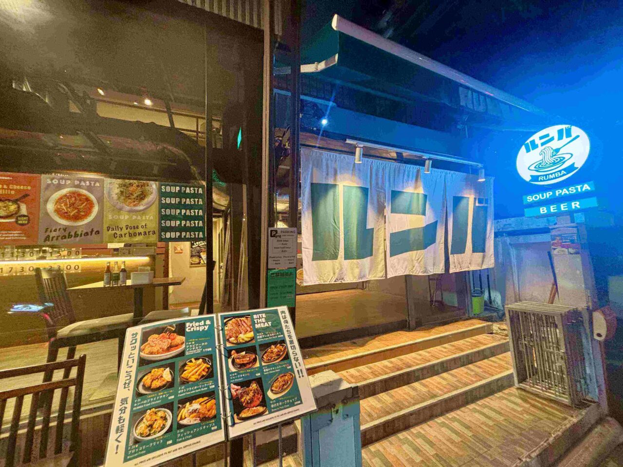A nighttime exterior photograph of Thonglor Rumba restaurant in Bangkok, featuring a large white fabric noren curtain with the word Rumba written in green Japanese katakana characters, a glowing circular blue sign, and an illustrated menu board displayed at the entrance steps. Reference filename: rumba-15.jpg.