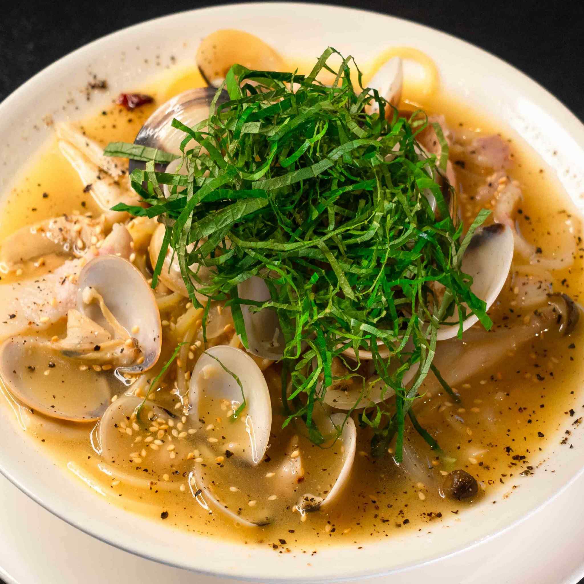 A close-up food photograph of Vongole Addiction, the signature Japanese-style clam soup pasta at Thonglor Rumba. The dish features springy noodles submerged in a rich, deeply savory clam broth, loaded with numerous fresh clams in their shells, shimeji mushrooms, and toasted sesame seeds, topped with a high, generous nest of finely shredded green shiso basil leaves. Reference filename: rumba-2.jpg.