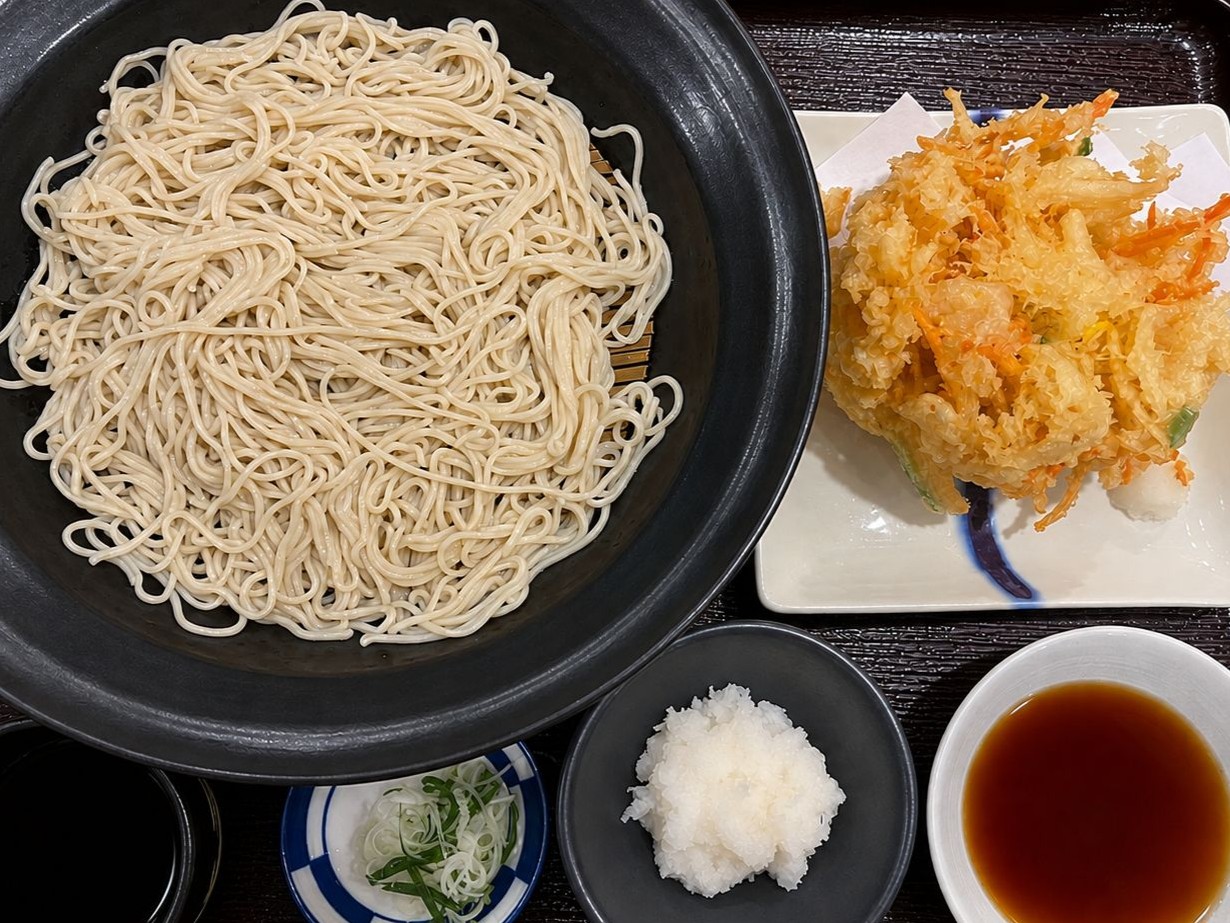 A food photograph of a signature Kakiage Tenmori cold soba set at Kisoba Azuma (Kisoba Azuma) Sukhumvit 49 in Thonglor, Bangkok. A large black bowl holds freshly made in-house cold soba noodles served alongside a massive golden-crisp mixed tempura fritter (Kakiage) with vegetables and shrimp, accompanied by soba dipping tsuyu broth, grated daikon radish, and chopped green onions. Reference filename: kisoba-azuma-skv49-2.jpg.