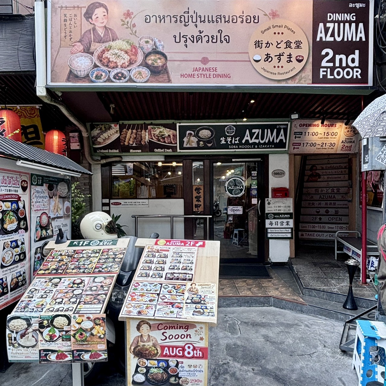 An exterior photograph showing the street-level entrance of Kisoba Azuma on Sukhumvit Soi 33/1 in Phrom Phong, Bangkok, with signs and menus pointing to Machikado Shokudo Azuma on the 2nd floor. Features outdoor menu stands, glowing red lanterns, and directional stairs. Reference filename: 13.jpg.