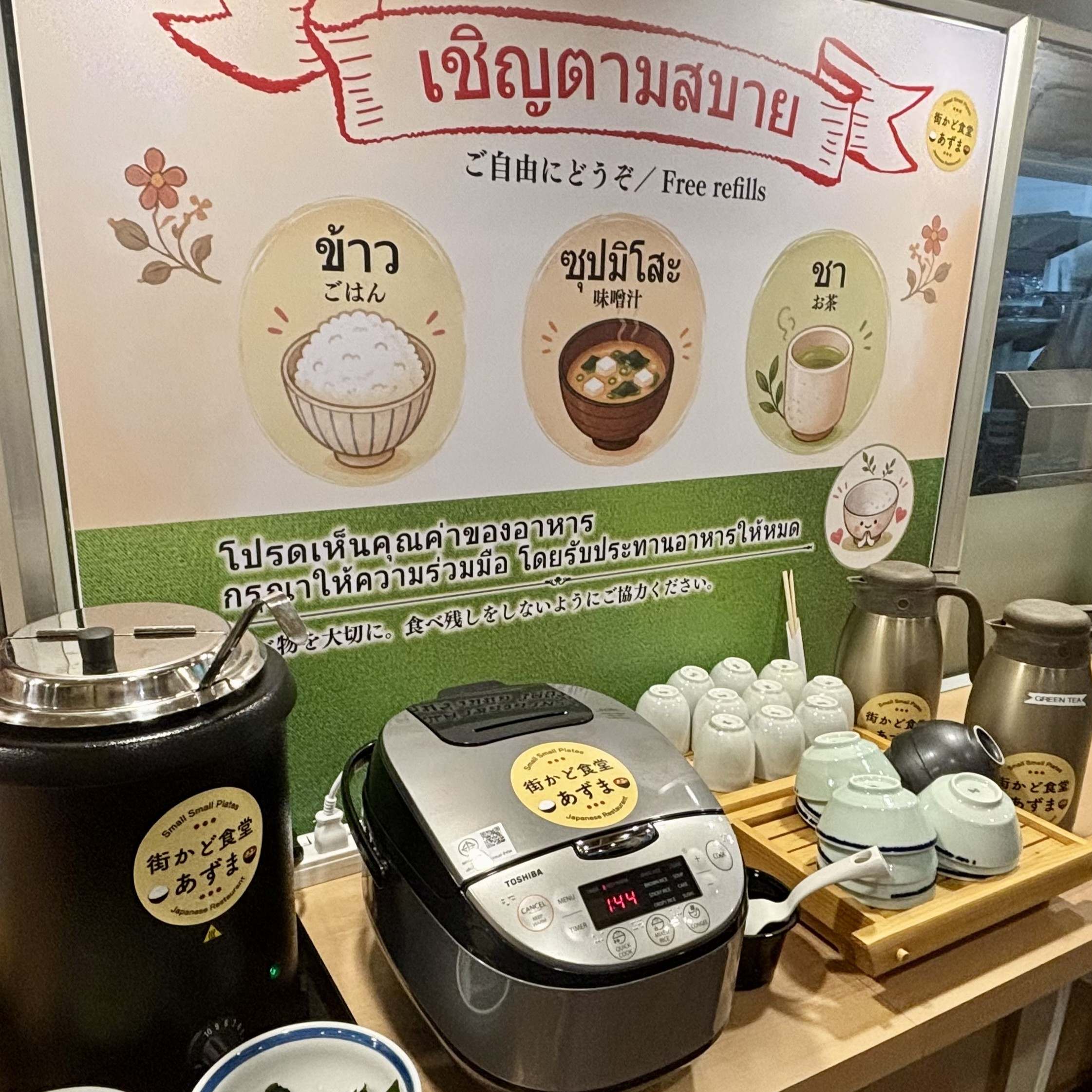 A facility photograph showcasing the self-service free refill station at Machikado Shokudo Azuma in Sukhumvit 33/1, Phrom Phong, Bangkok. A large electric rice cooker, a hot soup warmer for daily miso soup, tea thermoses, and stacked bowls are arranged neatly beneath a multilingual signboard in Japanese, Thai, and English stating "Free refills of rice, miso soup, and tea." Reference filename: machikado-shokudo-azuma-3.jpg.