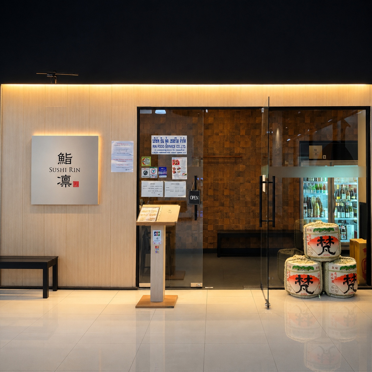 An exterior entrance view of 鮨凛 SUSHIRIN on the 4th floor of RSU タワー in Phrom Phong (Sukhumvit 31, バンコク. The modern Japanese facade features a light wood wall with the backlit Sushi Rin logo sign, glass entrance doors, a menu stand, stacked traditional Born sake barrels, and an illuminated sake refrigerator visible inside. Reference filename: sushi-rin-15.jpg.
