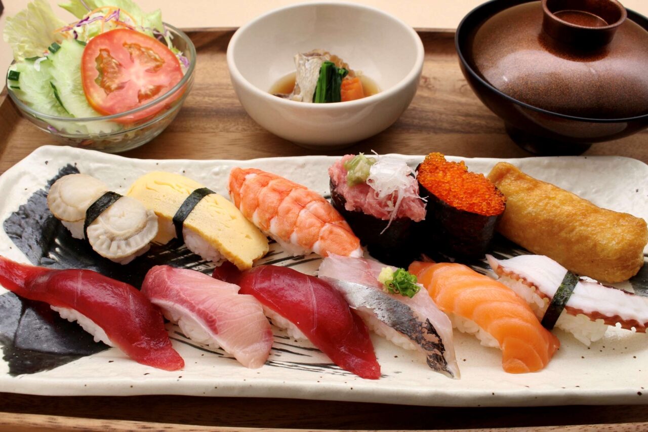 An authentic Japanese large nigiri sushi lunch set on a rectangular ceramic plate at Sushi Rin in Phrom Phong, Bangkok (520 THB). The platter features a generous assortment of 12 pieces of sushi including tuna (Akami), salmon, yellowtail, horse mackerel, boiled shrimp, scallop, octopus, tamago, inari, and gunkan maki, served on a wooden tray with a fresh garden salad, a simmered side dish, and miso soup. Reference filename: sushi-rin-3.jpg.