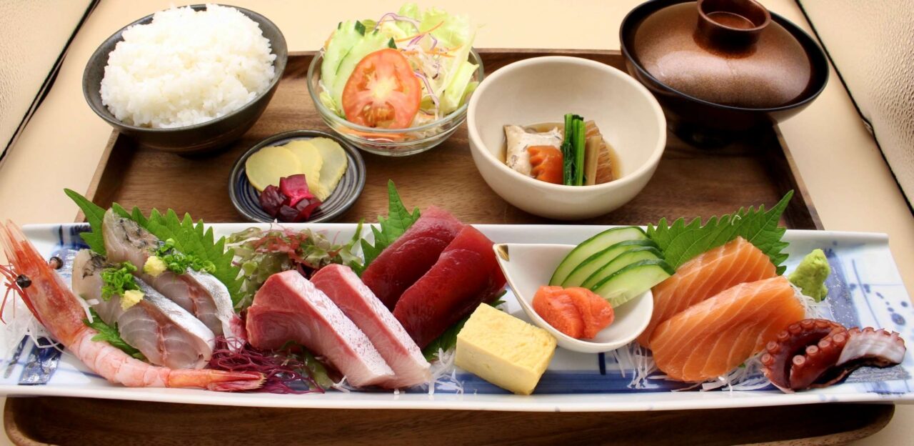 An authentic Japanese assorted sashimi lunch set on a rectangular ceramic plate at Sushi Rin in Phrom Phong, Bangkok (590 THB). The platter showcases a generous variety of fresh seafood including whole sweet shrimp, thick-cut yellowtail, lean tuna (Akami), salmon, octopus, marinated fish, seasoned cod roe (Mentaiko), and rolled omelet (Tamago), served on a wooden tray with steamed rice, fresh garden salad, a simmered Japanese side dish, pickles, and miso soup. Reference filename: sushi-rin-4.jpg.