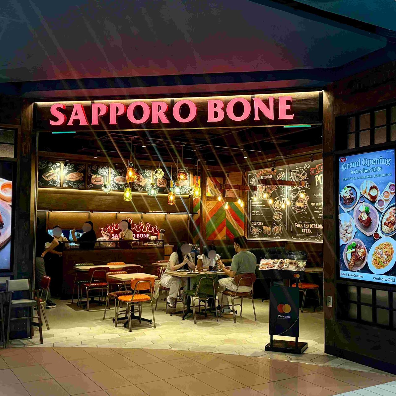 The storefront of SAPPORO BONE on the 6th floor of CentralWorld in Bangkok. The warm, wood-toned interior features cozy dining tables and customers enjoying their meals, while a digital display at the entrance highlights its grand opening on October 9, 2026, marking its first-ever branch in Thailand from Japan. File name: Sapporo-bone-15.jpg
