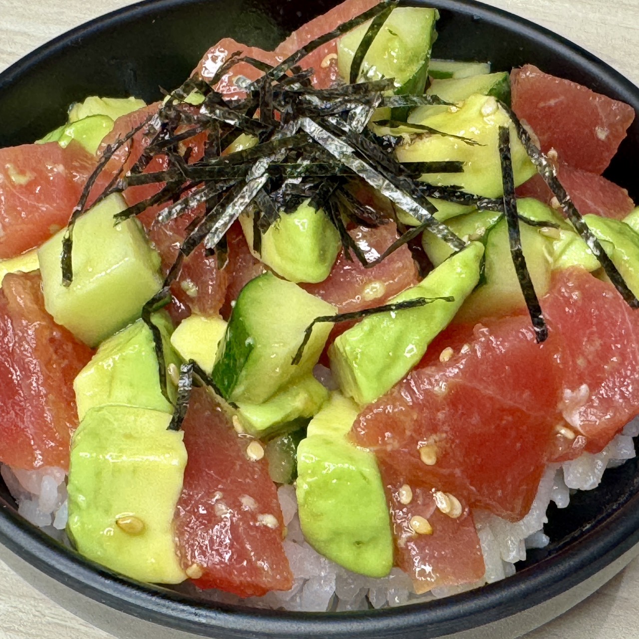 A close-up shot of a Japanese-style tuna poke bowl served in a black bowl at Kaisen Tengoku. The dish features fresh diced raw red tuna, avocado, and cucumber over white rice, garnished with toasted sesame seeds and shredded nori seaweed. Photographed by Kaori Kondo.