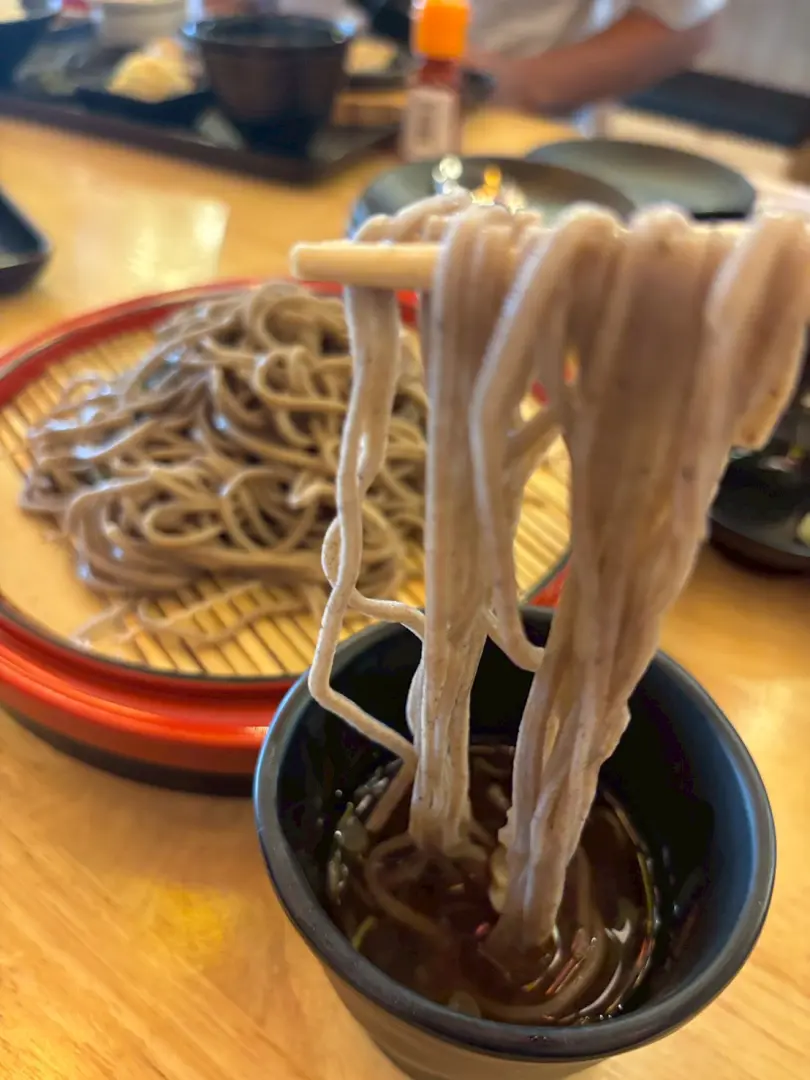 箸でつまんだざるそばを、そばつゆにつけようとしているところ。奥にはそばが盛られたざるが見える。