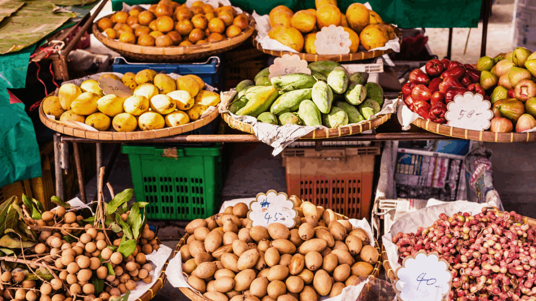 タイの市場に並ぶマンゴーなどの南国フルーツ。残留農薬のリスクがあるため、皮をむく果物でも食べる前には表面を洗うことが推奨されることを示す画像。