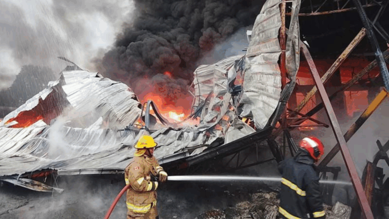 激しい炎と黒煙を上げながら燃え盛る工場の建物。手前には放水ホースを持ち、消火活動に当たる2人の消防士が写っている。
