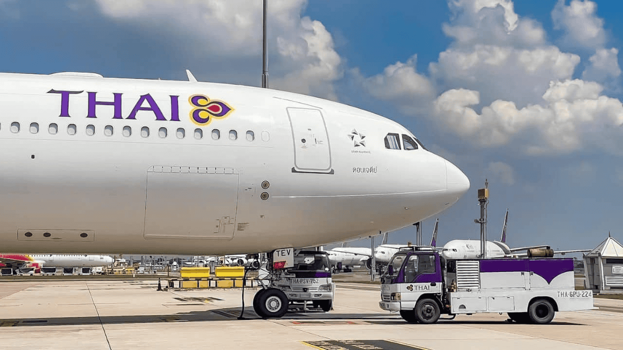 空港エプロンに駐機するタイ国際航空のワイドボディ機(白地に紫・金のTHAIロゴ)と紫白の地上サービス車両。青空と白い雲を背景に撮影。