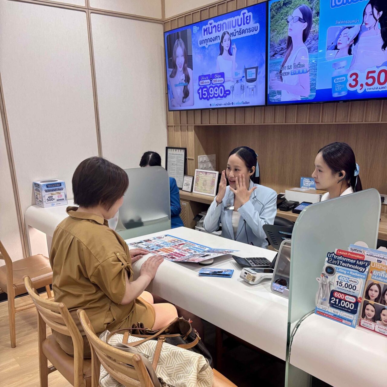 バンコクの美容クリニック「Aura Bangkok Clinic」のドンキモールトンロー店にて、スタッフがお客様に対して丁寧にカウンセリングや施術の説明を行っている様子を捉えた写真。手前のカウンターには各種プロモーションの卓上ポップが置かれ、背景のモニターには施術案内が映し出されています。ファイル名:14.jpg