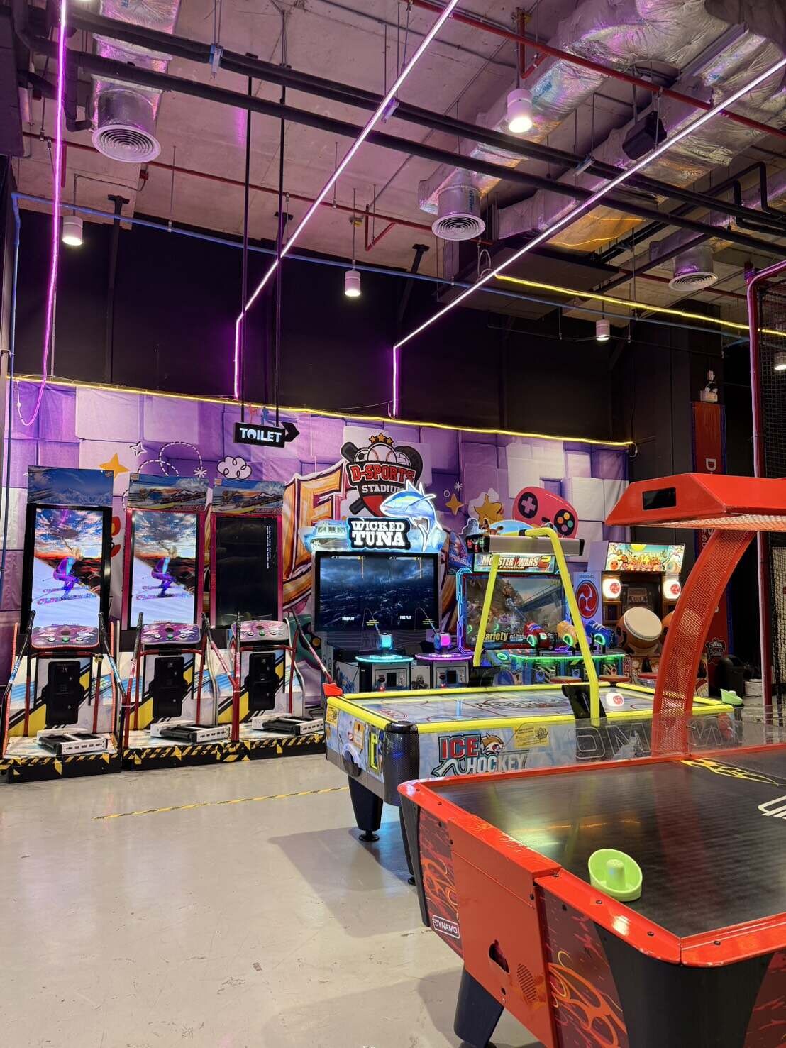バンコクのドンキモール・トンロー5階にある屋内スポーツ&エンターテインメント施設「D-Sports Stadium」のゲームエリアを撮影した写真。手前には黄色や赤のエアホッケー台が置かれ、奥にはスキーゲーム機や「Wicked Tuna」などのシューティング、アーケードゲーム機がカラフルなネオン照明のもとで並んでいます。ファイル名:15.jpg