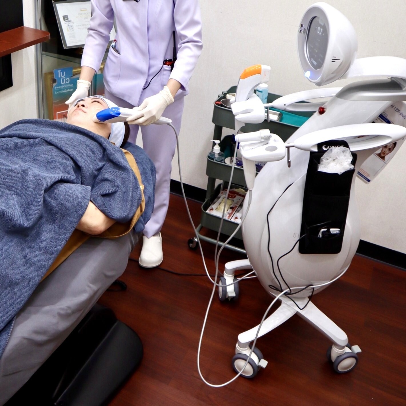 バンコクの美容クリニック「Aura Bangkok Clinic」の施術室にて、施術着を着てベッドに横たわる患者に対して、医療スタッフが専用の照射機器やハンドピースを使用してフェイシャルまたはリフトアップの施術を行っている様子を捉えた写真。ファイル名:15.jpg