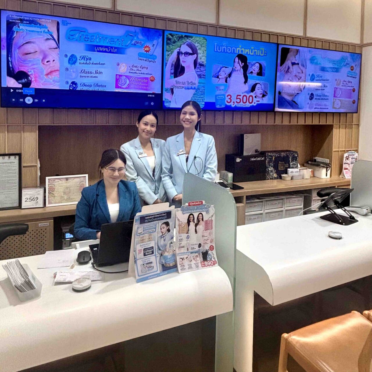 バンコクのドンキモール・トンロー2階にあるAura Bangkok Clinicの受付カウンターと、笑顔で迎えるスタッフの様子を撮影した写真。背景のモニターには、各種スキンケア施術やプロモーション情報のデジタルサイネージが表示されています。ファイル名:2.jpg