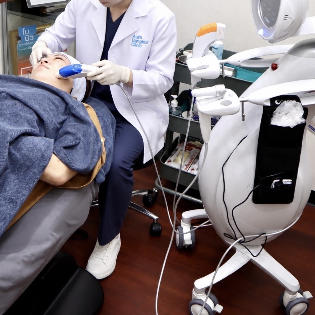 バンコクの美容クリニック「Aura Bangkok Clinic」の施術室にて、施術着を着てベッドに横たわる患者に対して、医療スタッフが専用の照射機器やハンドピースを使用してフェイシャルまたはリフトアップの施術を行っている様子を捉えた写真。ファイル名:15.jpg